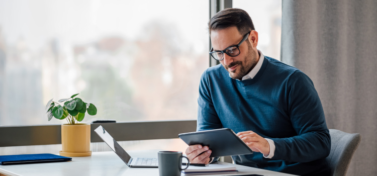 Picture of a man sitting at a desk looking his tablet in front of a laptop.