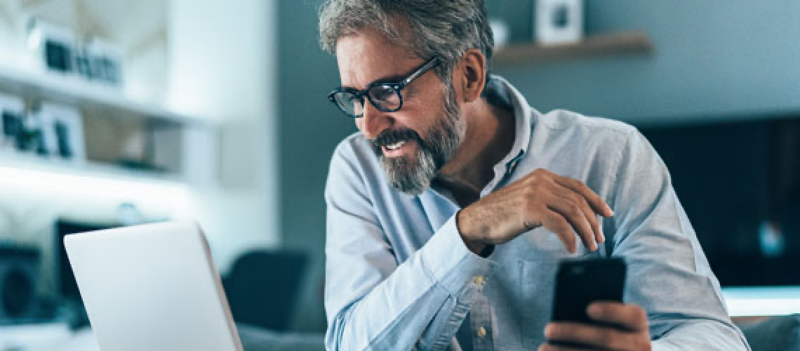 Photo of a man smiling while holding his phone and looking at his computer 
