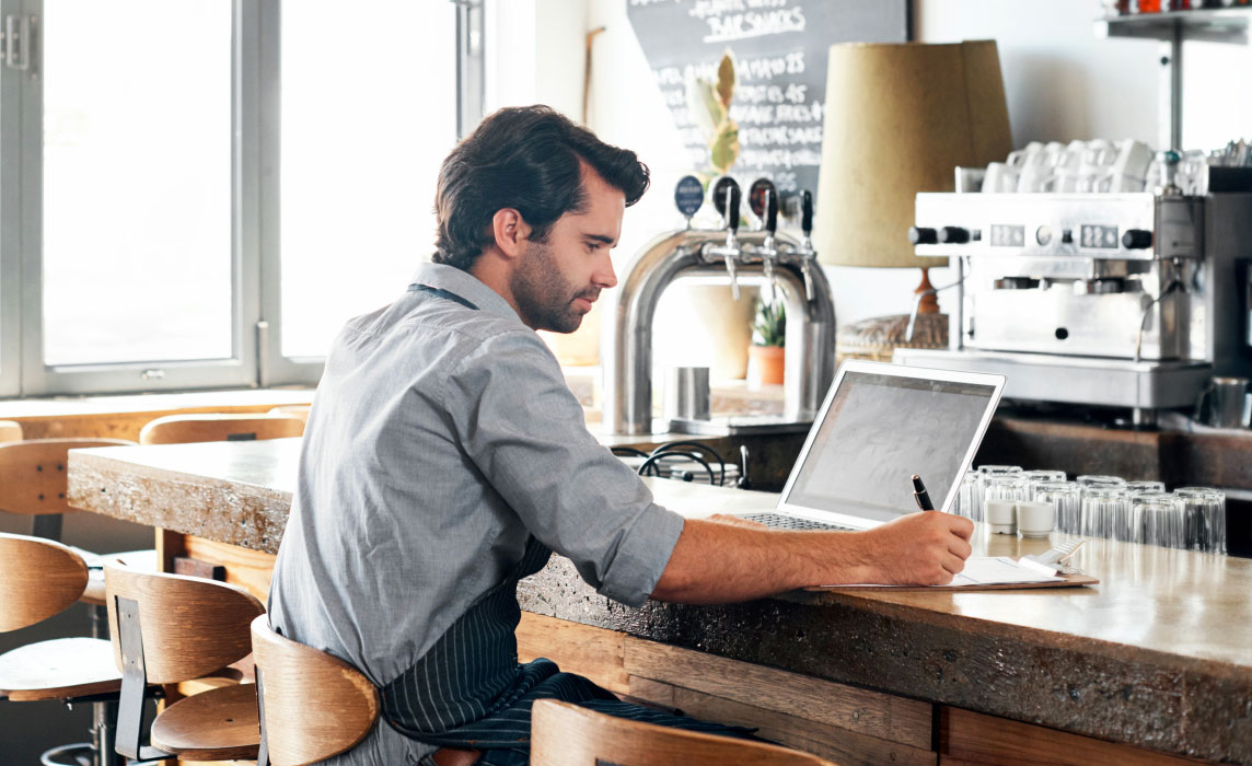 Photo of a business owner with an apron sitting at his bar during the day and working on his computer 